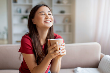 Happy Asian Woman Enjoying Cup Of Coffee, Relaxing On Couch At Home