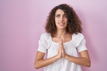 Hispanic woman with curly hair standing over pink background begging and praying with hands...