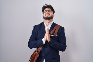 Hispanic man with beard wearing business clothes begging and praying with hands together with hope...