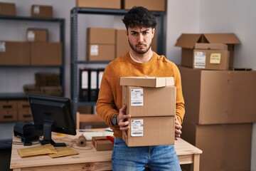 Hispanic man with beard working at small business ecommerce holding packages skeptic and nervous,...