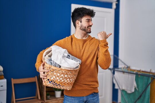Hispanic man with beard holding laundry basket at laundry room pointing thumb up to the side smiling happy with open mouth