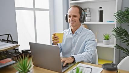 Fototapeta na wymiar Cheerful middle age man, a successful executive, joyfully working at his office desk, listening to music and sipping his morning espresso coffee, brightening up the indoor workplace.
