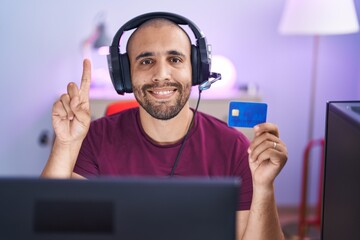 Hispanic man with beard doing online shopping with computer and credit card smiling with an idea or question pointing finger with happy face, number one