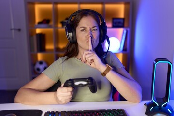 Beautiful brunette woman playing video games wearing headphones asking to be quiet with finger on...