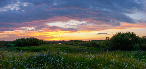 A wonderful sunset with bright clouds over a flowering meadow by the river. Tourism, outdoor...
