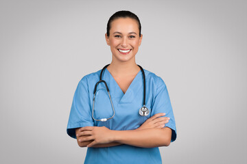 Portrait of positive european woman nurse posing with crossed arms and smiling at camera over grey...