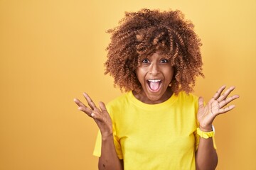 Young hispanic woman with curly hair standing over yellow background celebrating crazy and amazed...