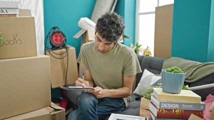 Young hispanic man writing on document sitting on sofa at new home