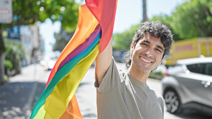 Young hispanic man smiling confident holding rainbow flag at street