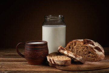 fresh bread sliced on a cutting board with a glass jar and a clay mug with milk on a brown background with a gradient. simple rustic still life. side view
