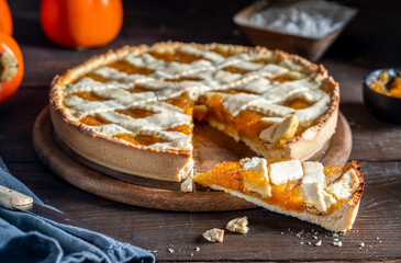 Italian homemade persimmon tart or crostata with one piece aside, ingredients over wooden background