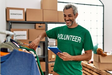 Middle age grey-haired man volunteer using touchpad holding clothes at charity center