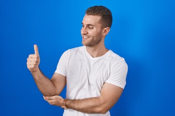 Fototapeta premium Young caucasian man standing over blue background looking proud, smiling doing thumbs up gesture to the side