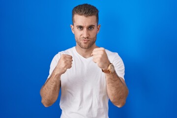 Young caucasian man standing over blue background ready to fight with fist defense gesture, angry and upset face, afraid of problem