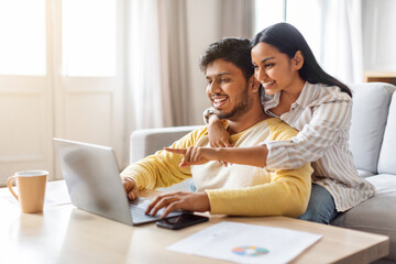 Happy young indian couple websurfing on laptop together at home - obrazy, fototapety, plakaty
