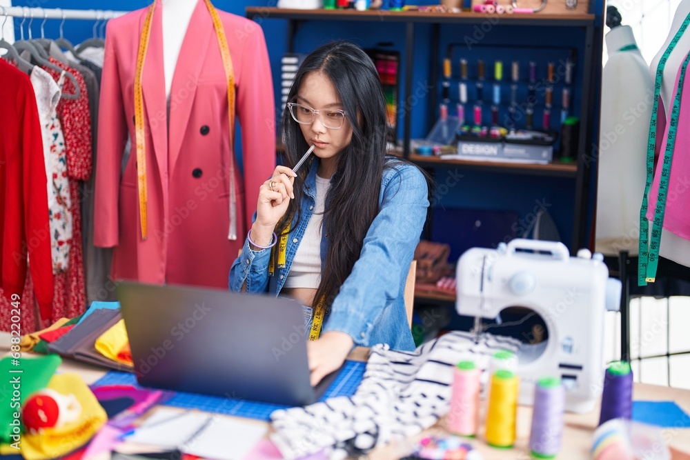 Poster Young chinese woman tailor using laptop with doubt expression at clothing factory