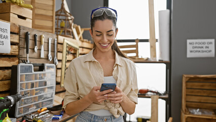 Smiling young hispanic woman, a beautiful carpenter, confidently texting on her smartphone amid the timber at an indoor carpentry workshop.