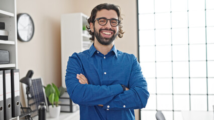 Young hispanic man business worker standing with arms crossed gesture smiling at the office
