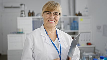 Confident middle age blonde woman scientist, smiling broadly, holding clipboard in bustling lab,...