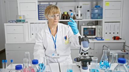 Serious, blonde middle-age woman scientist, taking notes and holding a test tube in a bustling lab,...
