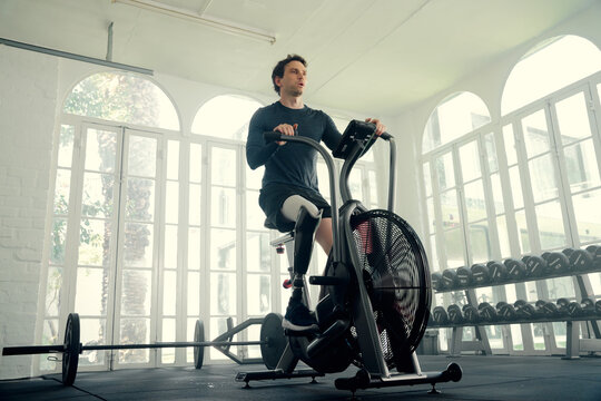 Young caucasian man in sports clothing cycling with prosthetic leg on exercise bike at the gym