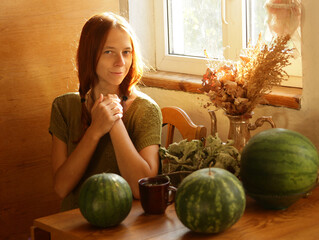 Fototapeta premium Selfie portrait of fair-haired woman in ethnic natural clothes in a country house, in the rays of the sun. What is sitting at the table on which there are large green watermelons.