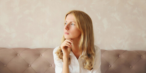 Pensive young woman thinks looking away while sitting on the couch in living room at home