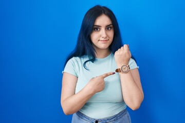 Young modern girl with blue hair standing over blue background in hurry pointing to watch time, impatience, looking at the camera with relaxed expression