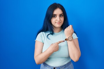 Young modern girl with blue hair standing over blue background in hurry pointing to watch time,...