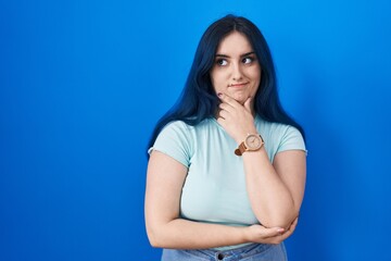 Young modern girl with blue hair standing over blue background thinking worried about a question, concerned and nervous with hand on chin