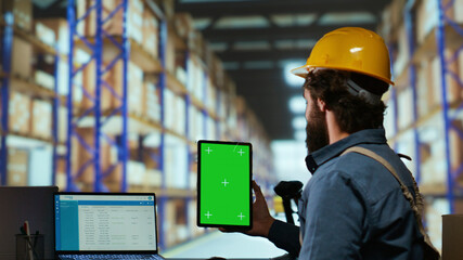 Warehouse operator holds greenscreen on tablet before working on merchandise distribution, examining isolated display on mobile device. Industrial staff member looks at blank chromakey screen.