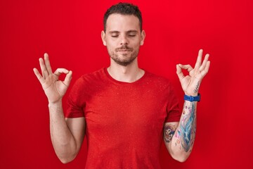 Fototapeta premium Young hispanic man standing over red background relaxed and smiling with eyes closed doing meditation gesture with fingers. yoga concept.