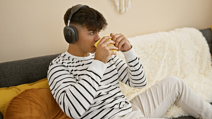 Obraz premium Attractive young hispanic man seriously engrossed in listening to music via headphones, relaxed on his living room sofa, savoring his morning espresso coffee at home