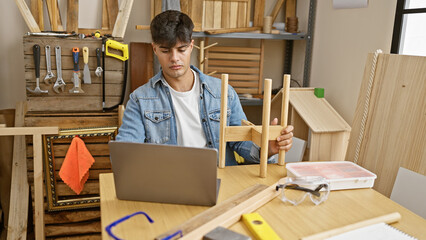 Handsome young hispanic carpenter working on a wood object at carpentry workshop, adeptly...