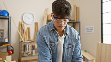 Handsome, young hispanic man standing tall in carpentry work studio â€“ a portrait of dedication and seriousness. this glasses-clad, professional carpenter is a master in the realm of woodworking - obrazy, fototapety, plakaty