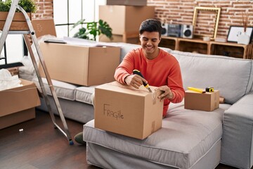 Young hispanic man smiling confident unpacking cardboard box at new home
