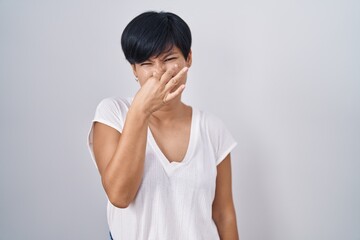 Young asian woman with short hair standing over isolated background smelling something stinky and disgusting, intolerable smell, holding breath with fingers on nose. bad smell