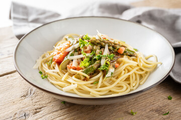 Spaghetti with vegetable sauce of green asparagus and tomatoes with parmesan and parsley garnish in a plate on a rustic wooden table, selected focus