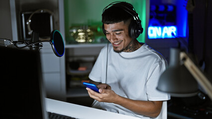 Smiling young latin man, a music studio maestro, happily listens to his phone's tunes, immersing in...