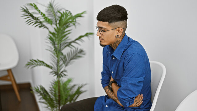 Suffering from stomachache, young hispanic man in business attire, tattooed and glasses-clad, sits in chair, nervous and in pain, awaiting interview in waiting room