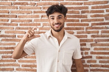 Arab man with beard standing over bricks wall background smiling and confident gesturing with hand doing small size sign with fingers looking and the camera. measure concept.