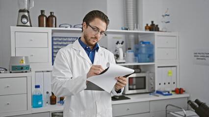 Young hispanic man scientist writing report standing at laboratory - obrazy, fototapety, plakaty