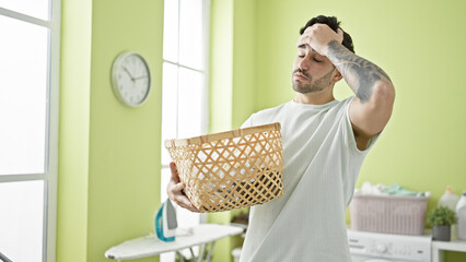 Young hispanic man holding basket with clothes stressed at laundry room