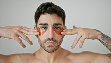 Young hispanic man standing shirtless putting baggy eyes patches over isolated white background