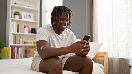 African american man using smartphone sitting on bed smiling at bedroom