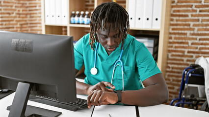 African american man doctor using computer looking watch with serious face at the clinic