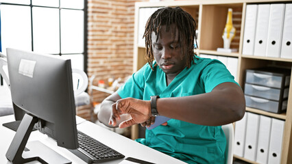 African american man doctor using computer looking watch with serious face at the clinic