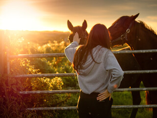 Teenager girl with long hair by a metal gate to a field with dark horses. Selective focus. Models...