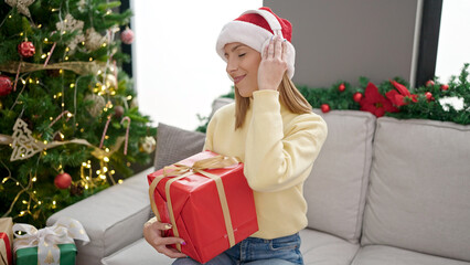 Obraz na płótnie Canvas Young blonde woman listening to music holding gift by christmas tree at home