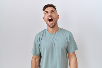 Hispanic man with beard standing over white background angry and mad screaming frustrated and...